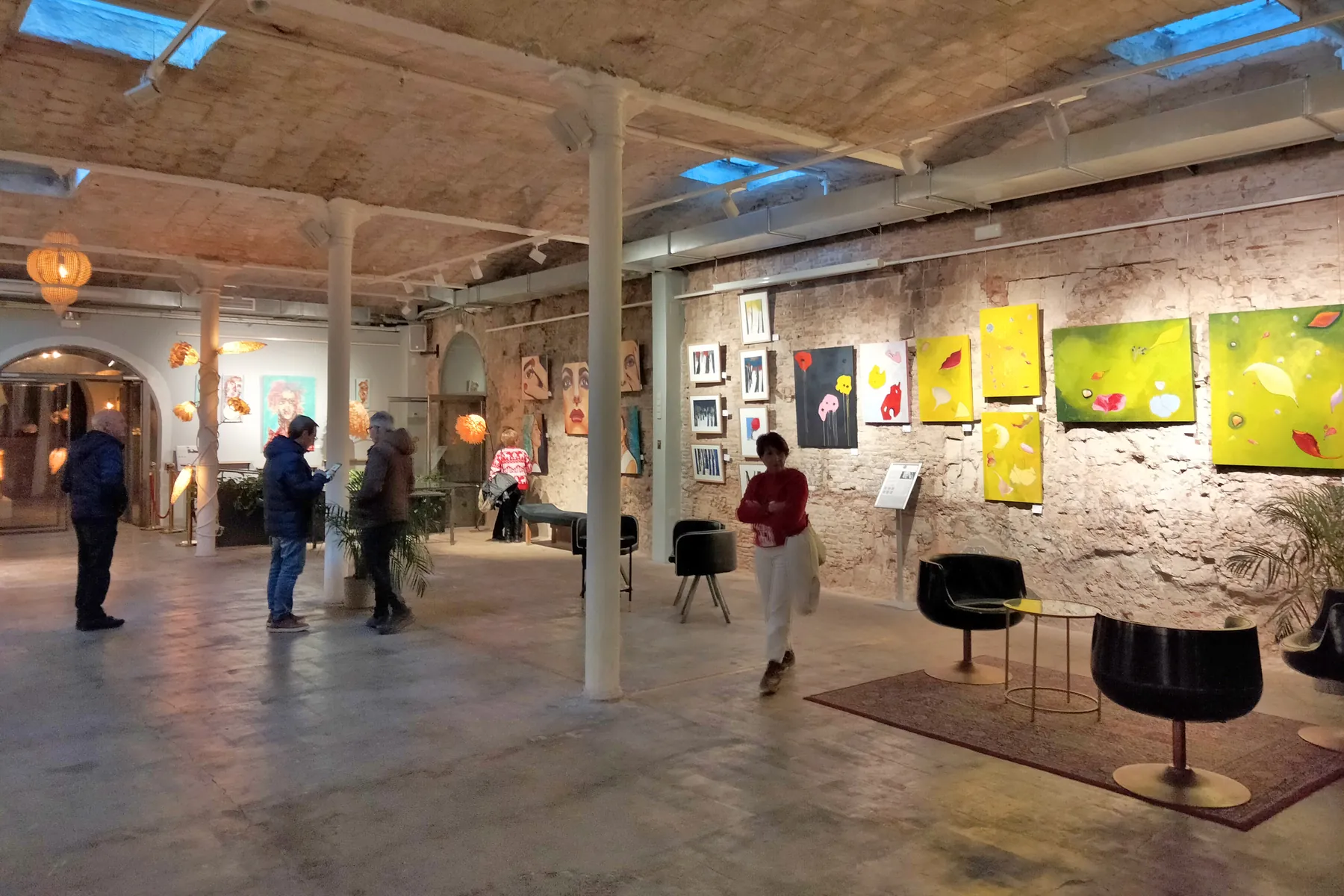 Visitors looking at paintings on the walls of an art gallery room with a vaulted ceiling and skylights